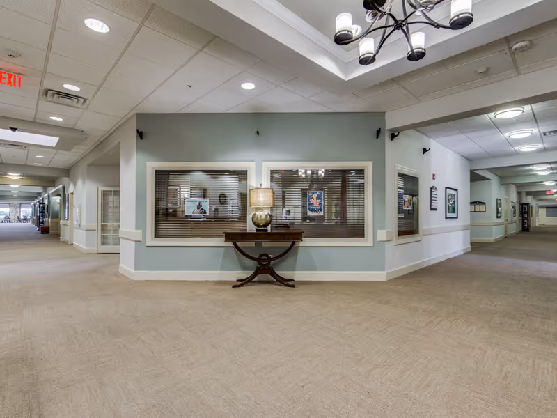 Interior hallway of a senior living facility with beige carpet and light blue walls. There is a wooden table with a lamp on it positioned against a wall with three large windows that have blinds. The hallway extends in two directions with framed pictures on the walls and ceiling lights. An exit sign is visible on the ceiling to the left.