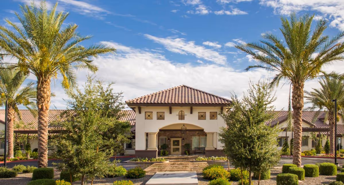 Front exterior view of Amber Creek Inn Memory Care Community building with a tiled roof, surrounded by palm trees and landscaped bushes under a partly cloudy blue sky.