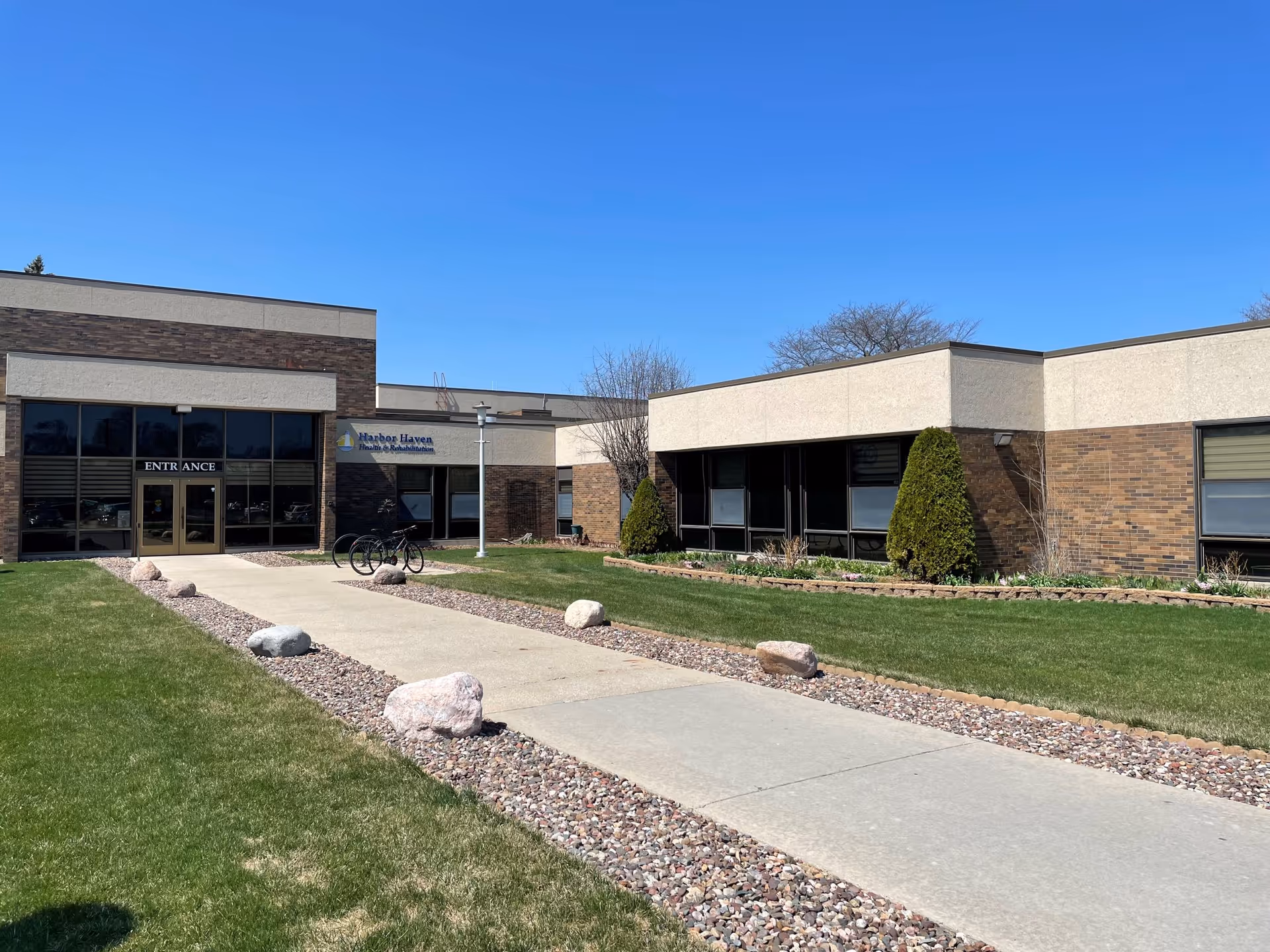 Exterior view of Harbor Haven Health & Rehabilitation building with a clear blue sky. The building has a brick and beige facade with large windows. A concrete walkway lined with rocks leads to the entrance, where a bicycle is parked near the door. There is green grass and some small shrubs around the building.