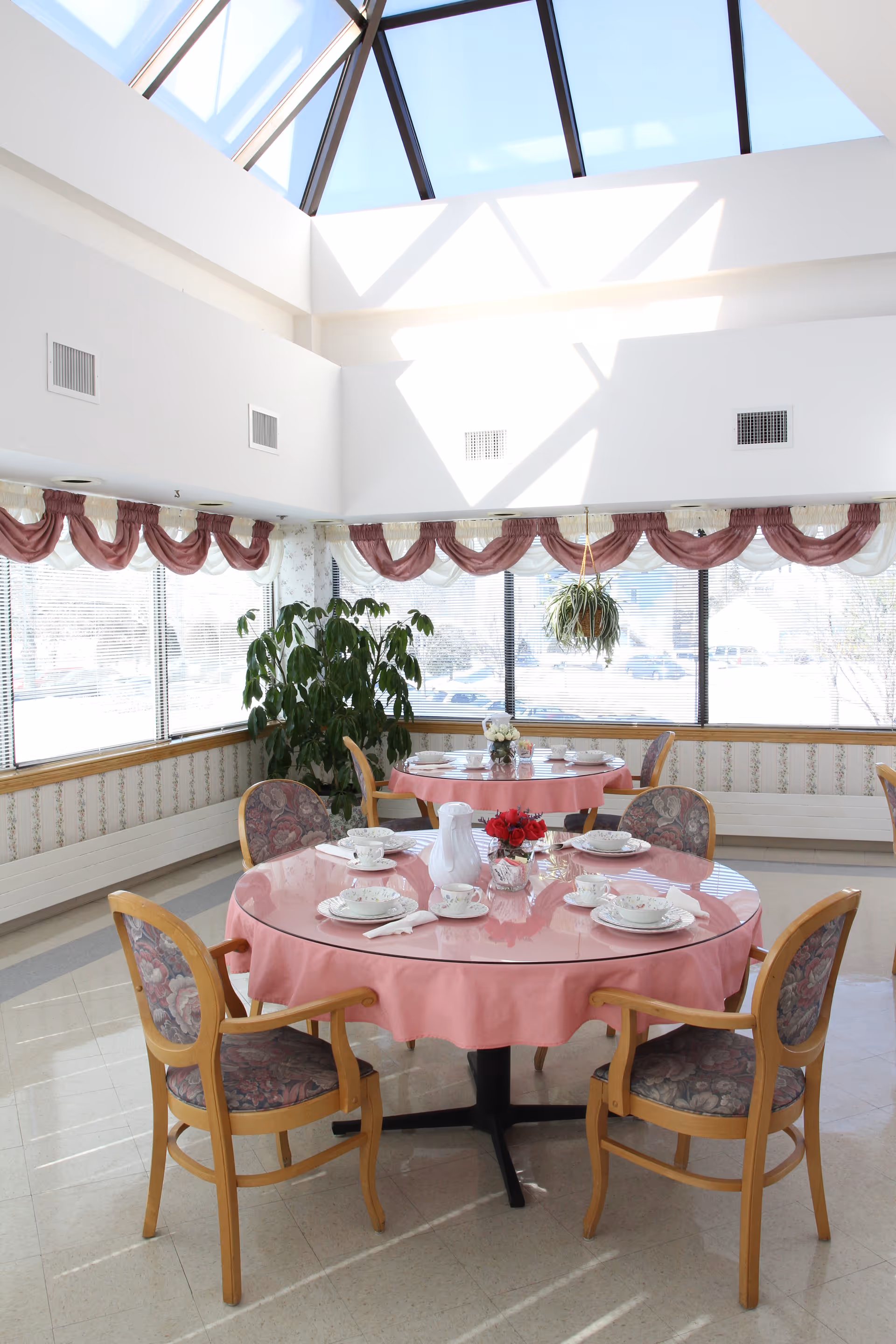 A bright dining room with large windows and a skylight ceiling. There are round tables covered with pink tablecloths, set with white china, cups, and a white pitcher. Four wooden chairs with floral upholstery surround each table. A green potted plant and a hanging plant are visible near the windows.