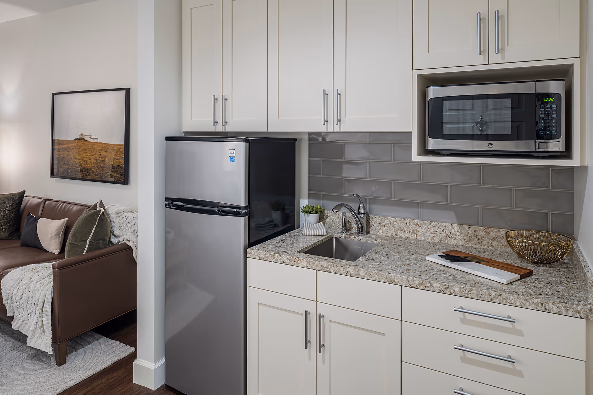 A modern kitchenette with white cabinets, a small stainless steel refrigerator, a microwave oven, a granite countertop with a sink, a cutting board, and a decorative bowl. To the left, part of a living room is visible with a brown leather sofa, cushions, a throw blanket, and a framed picture on the wall.