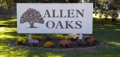 A large Allen Oaks sign with a tree logo sits on a flower-filled lawn.