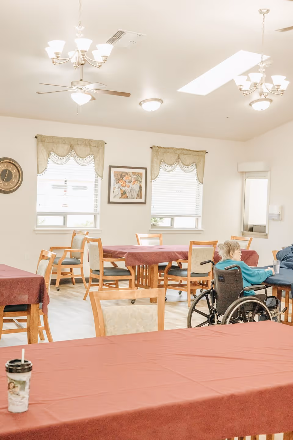 A bright dining room with several wooden tables covered with maroon tablecloths and matching chairs. Two windows with decorative valances and blinds are on the far wall, with a framed painting between them. Ceiling lights and fans illuminate the room. An elderly person in a wheelchair is seated at one of the tables.