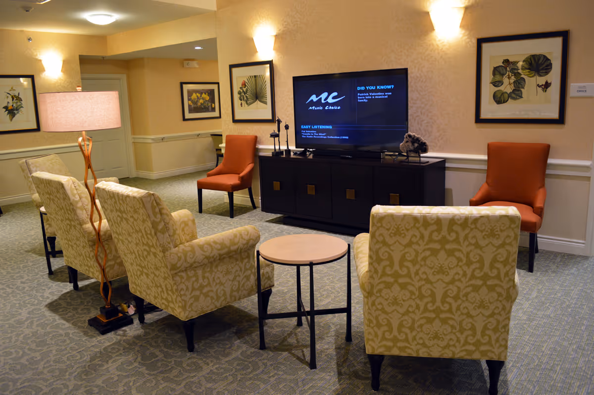 Comfortable senior living lounge with patterned armchairs and orange accent chairs arranged around a TV on a media console.