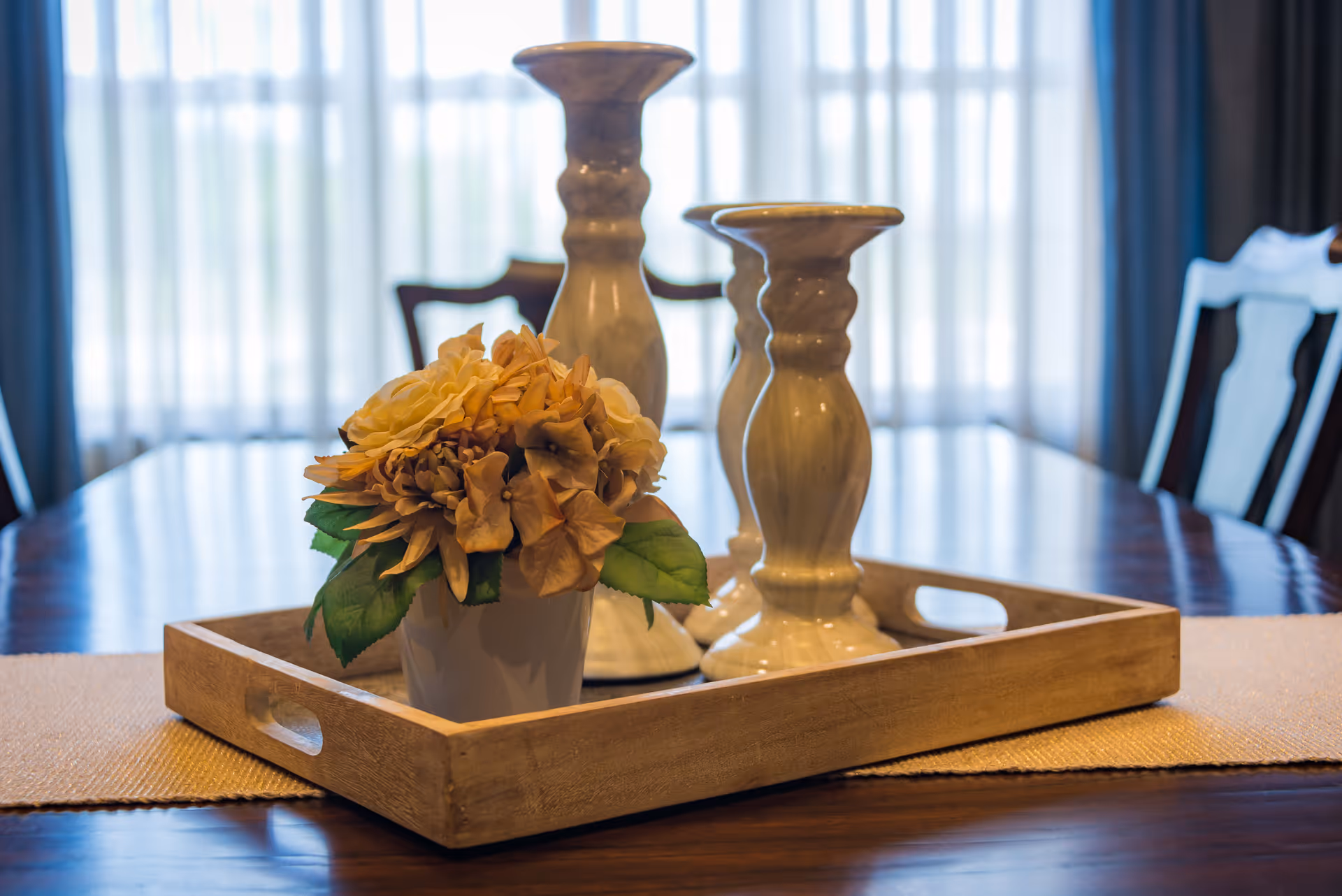 A wooden tray on a polished wooden dining table holding a small white pot with artificial yellow flowers and two beige ceramic candlesticks. The background shows sheer white curtains with dark drapes and wooden dining chairs.