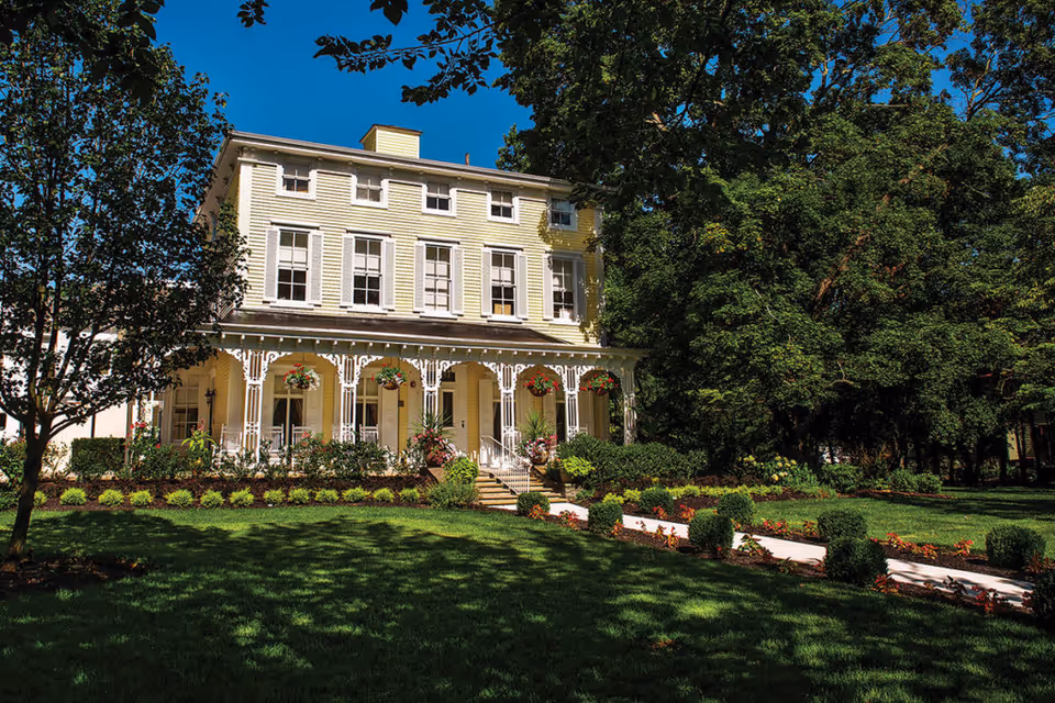 A three-story yellow Victorian-style building with a decorative front porch, manicured lawn, and large trees.
