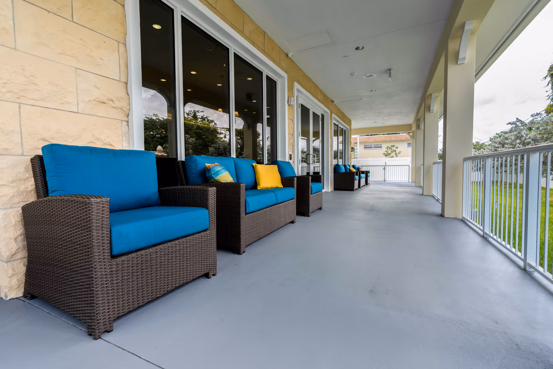 Covered outdoor porch with wicker lounge chairs featuring bright blue cushions lined along a building exterior.
