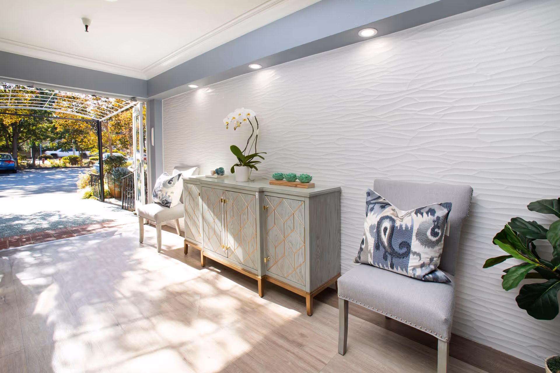 Bright modern interior entry/lounge with two upholstered chairs, a decorative console, textured white wall and open view to the driveway.