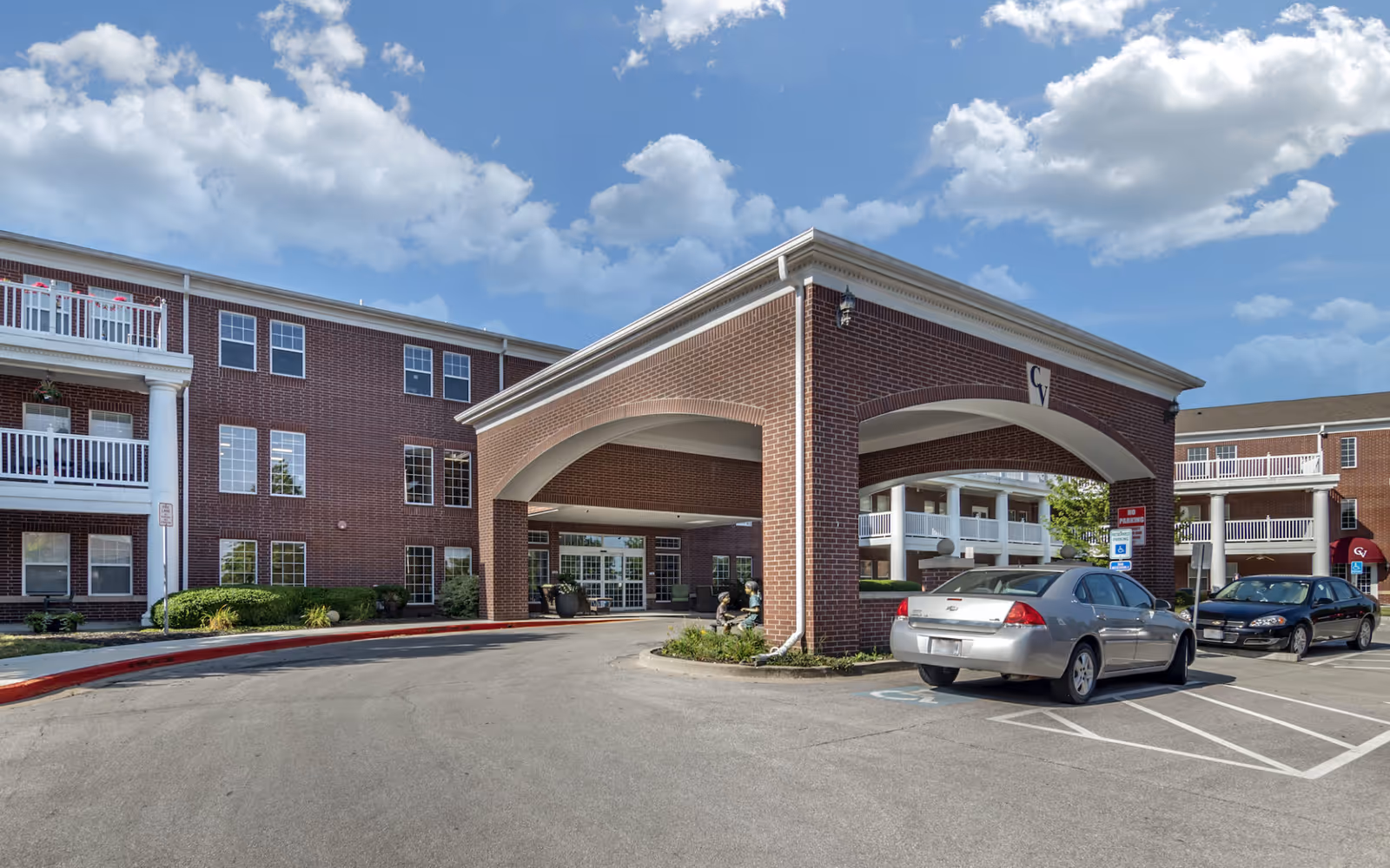 Exterior view of Carnegie Village Rehabilitation & Health Care Center showing a brick building with a covered entrance driveway, parked cars, and a clear blue sky with some clouds.