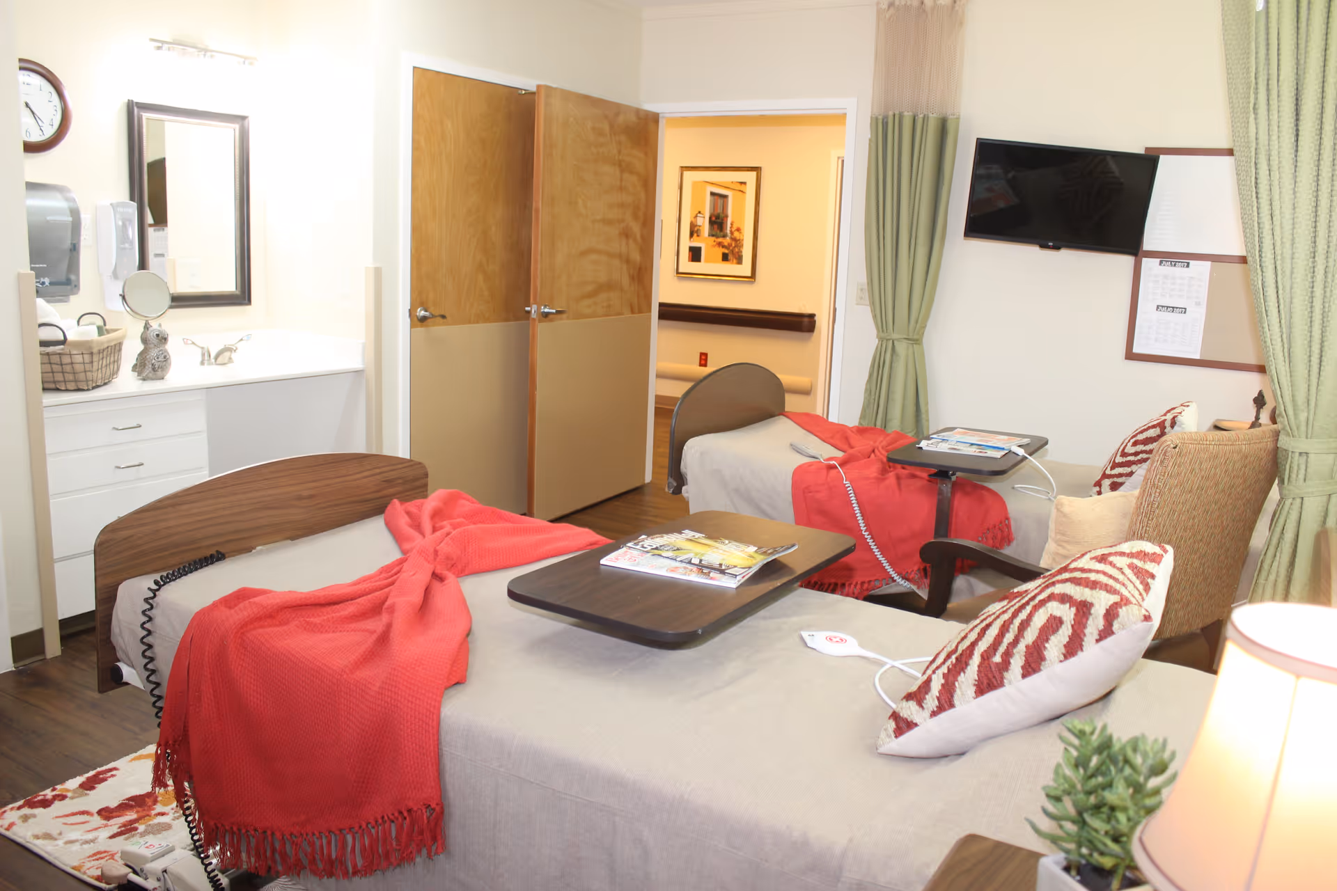 A nursing facility room with two beds, each covered with beige sheets and red blankets. Each bed has a tray table with magazines on it. There is a chair with patterned pillows, a wall-mounted TV, and a small sink area with a mirror and soap dispenser. The room has wooden doors and green curtains.