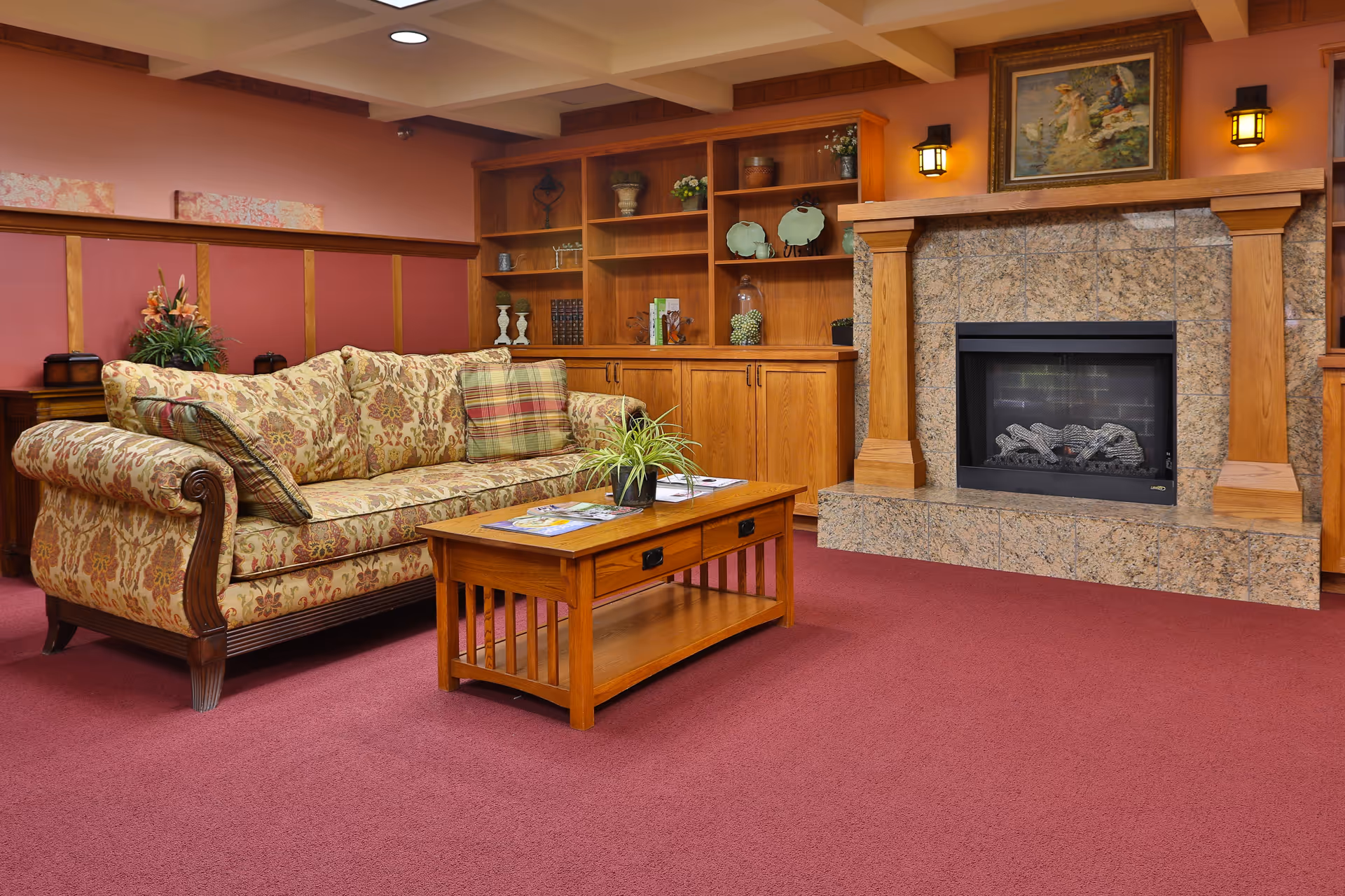 A cozy living room with a floral patterned sofa, a wooden coffee table with a small plant and magazines, a built-in wooden bookshelf with decorative items, and a stone fireplace with a painting above it. The room has a red carpet and warm lighting.