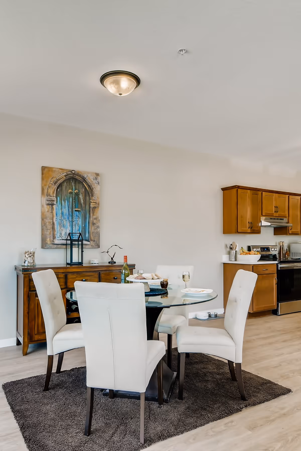 Glass-top dining table with four upholstered chairs on a rug in an open dining area next to a kitchen and sideboard with wall art.