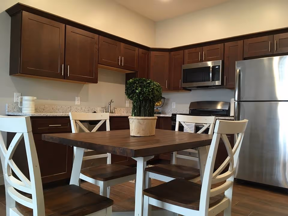 A modern kitchen with dark wood cabinets, a stainless steel refrigerator and microwave, a stove, and a wooden dining table with four white chairs. A small potted plant is placed in the center of the table.