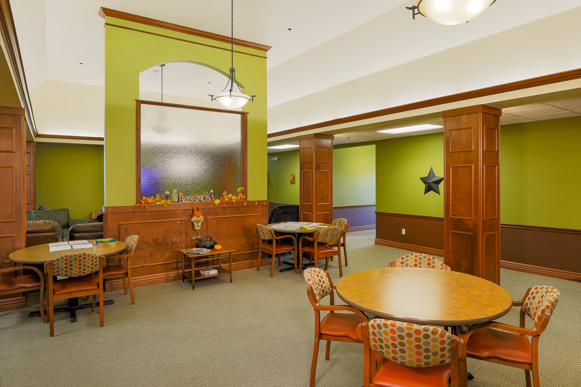 Interior view of a senior living facility common area with round tables and chairs featuring patterned upholstery. The walls are painted green with wooden paneling and columns. There is a decorative glass partition with autumn-themed decorations and a sign that reads 'Blessings'. Comfortable seating is visible in the background.