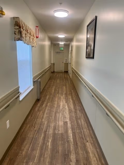 A long, narrow hallway with wood-patterned flooring and beige walls. There are handrails on both sides of the hallway, a window with a floral valance on the left wall, a framed picture on the right wall, and ceiling lights evenly spaced along the corridor. An exit sign and door are visible at the far end of the hallway.