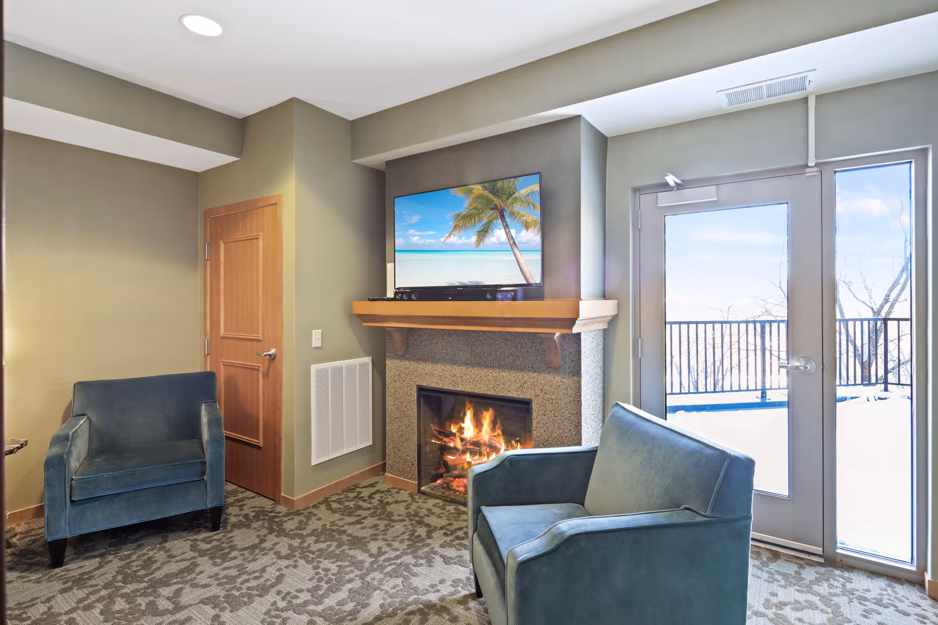 Small sitting room with two blue armchairs, a lit fireplace, a wall-mounted TV, and a glass door leading to a snowy balcony.