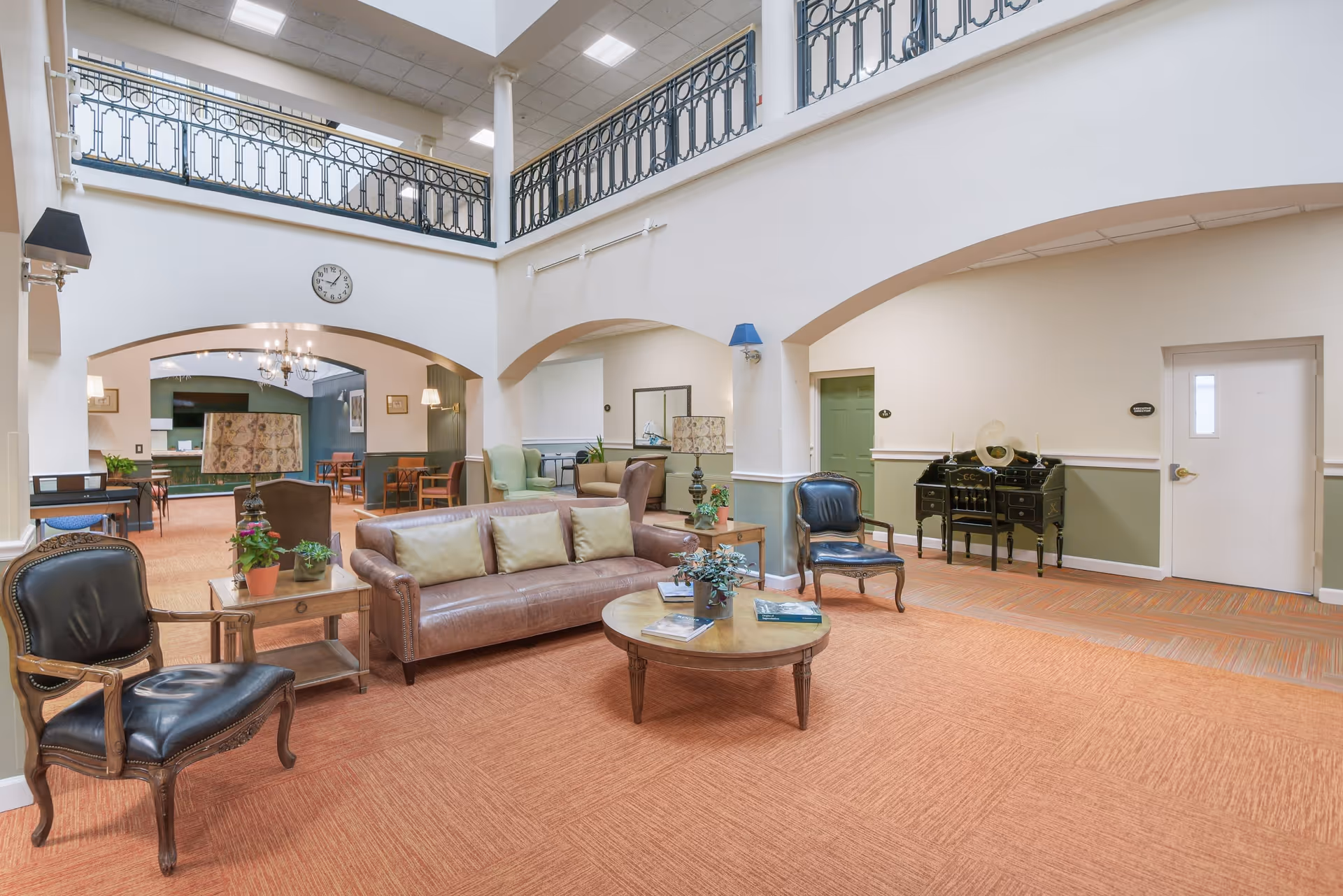 A spacious senior living community common area with a high ceiling and a second-floor balcony railing. The room features a brown leather sofa with three cushions, two black leather armchairs, a round wooden coffee table with books and plants, and a side table with a lamp and plants. In the background, there are additional seating areas with chairs and tables, a chandelier, and a clock on the wall.