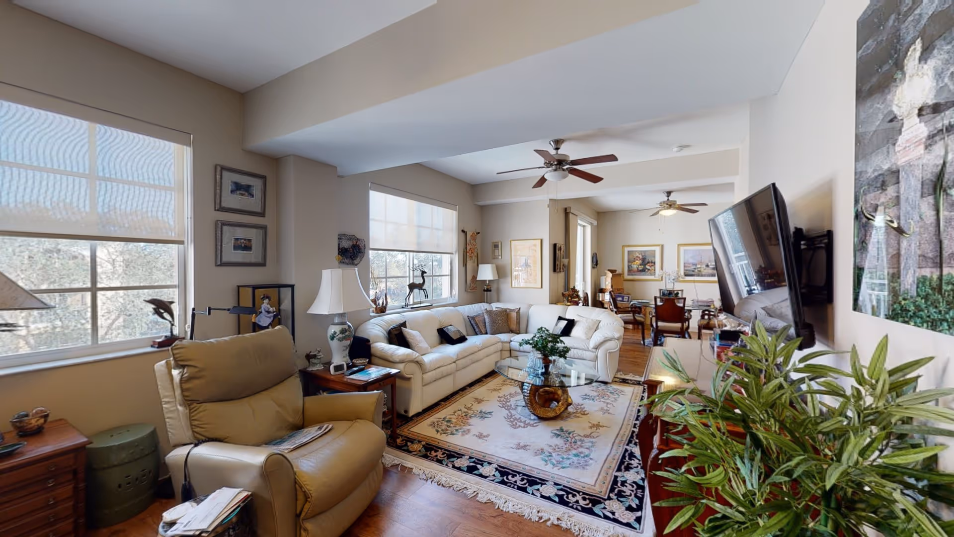 A bright and spacious living room with large windows covered by light roller shades. The room features a beige leather recliner, a white sectional sofa with multiple cushions, a glass coffee table on a floral area rug, and ceiling fans. There are framed pictures on the walls, a flat-screen TV mounted on the wall, and various decorative items including plants and lamps. In the background, a dining area with a wooden table and chairs is visible.