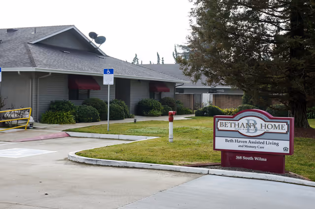 Exterior view of Bethany Home Society - Assisted Living (Beth Haven) showing a single-story building with a gray roof, bushes along the walls, a handicapped parking sign, and a sign in the grass that reads Bethany Home, Beth Haven Assisted Living and Memory Care, 368 South Wilma.
