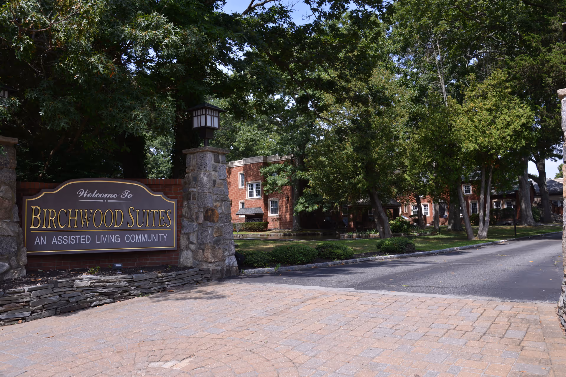 Entrance to Birchwood Suites, an assisted living community, featuring a stone and brick sign with lanterns on stone pillars, surrounded by trees and greenery, with a paved driveway leading into the property where red brick buildings are visible in the background.