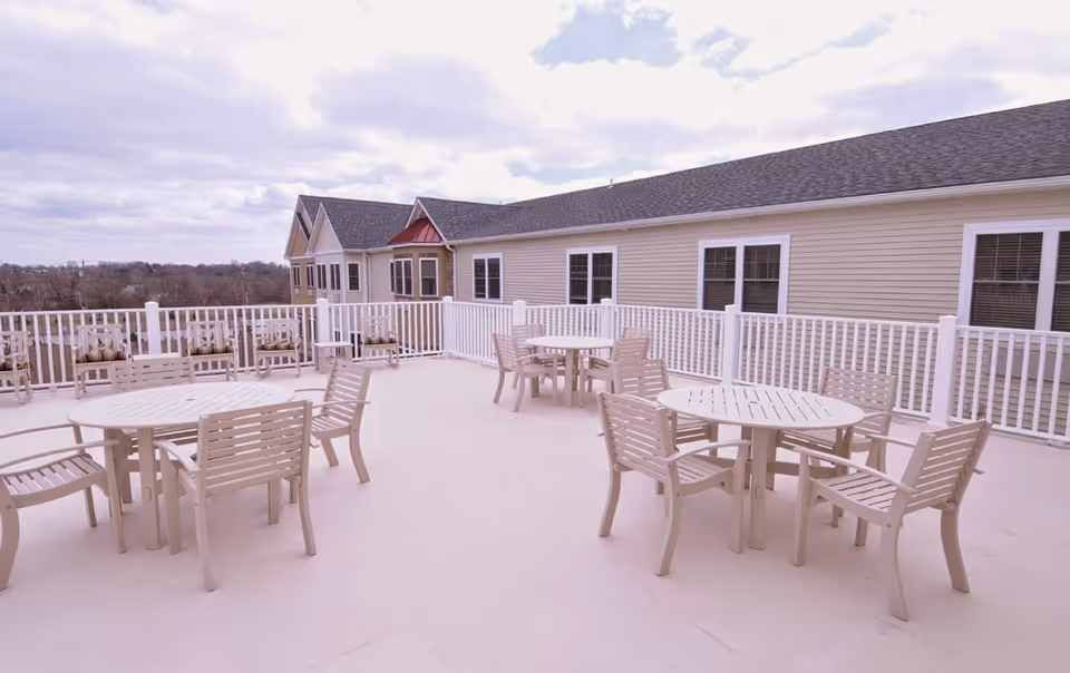 Rooftop patio with multiple round tables and chairs and a white railing beside a beige senior living building.
