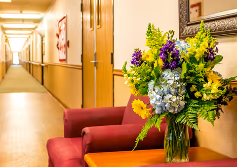 A long, well-lit hallway in a residential facility with wooden doors along the walls. In the foreground, there is a red armchair next to a wooden table holding a large bouquet of colorful flowers including yellow, purple, and blue blooms. A framed mirror hangs on the wall above the table.