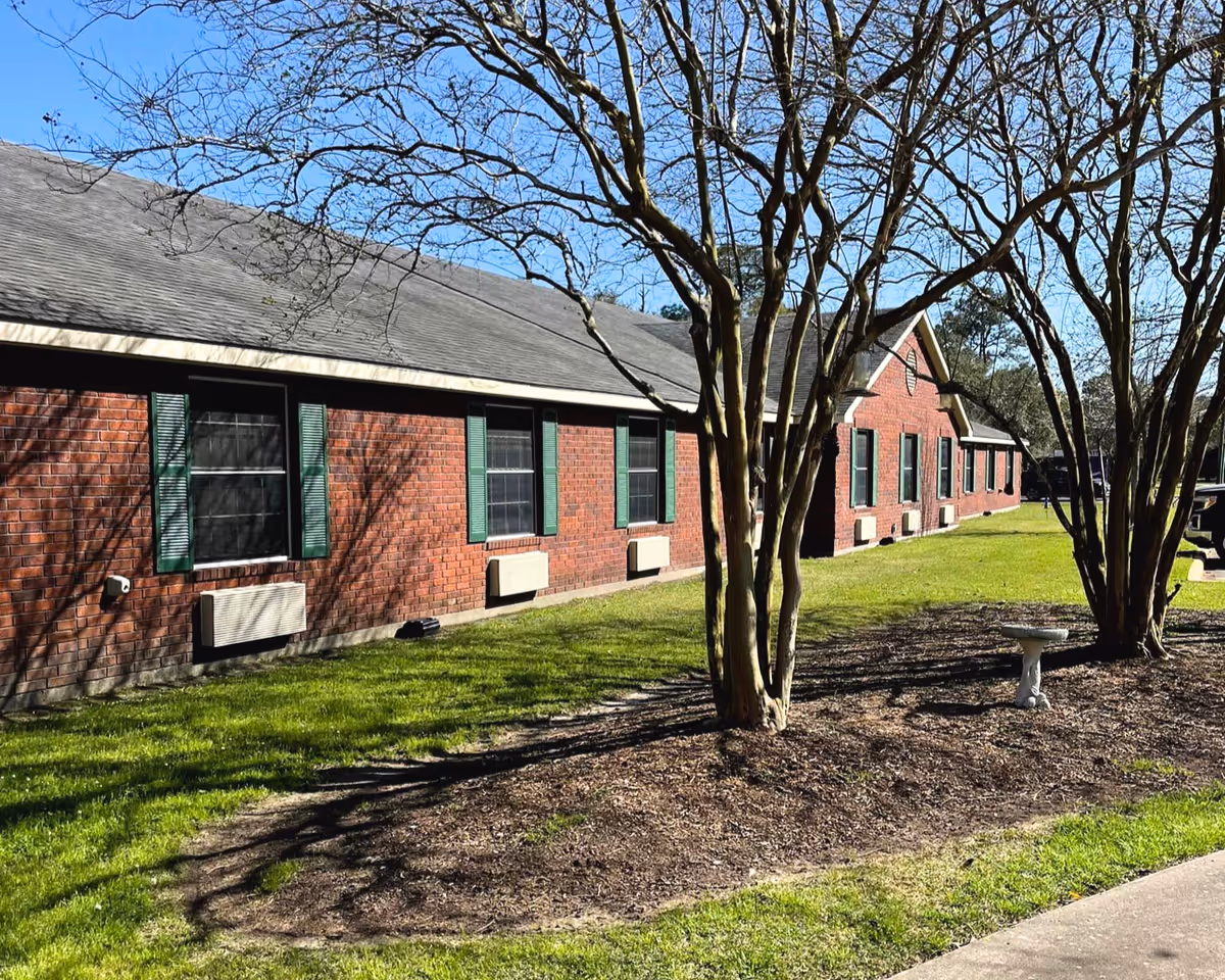 Single-story red brick nursing facility with green shutters, trees, a grassy lawn and a small birdbath in front.
