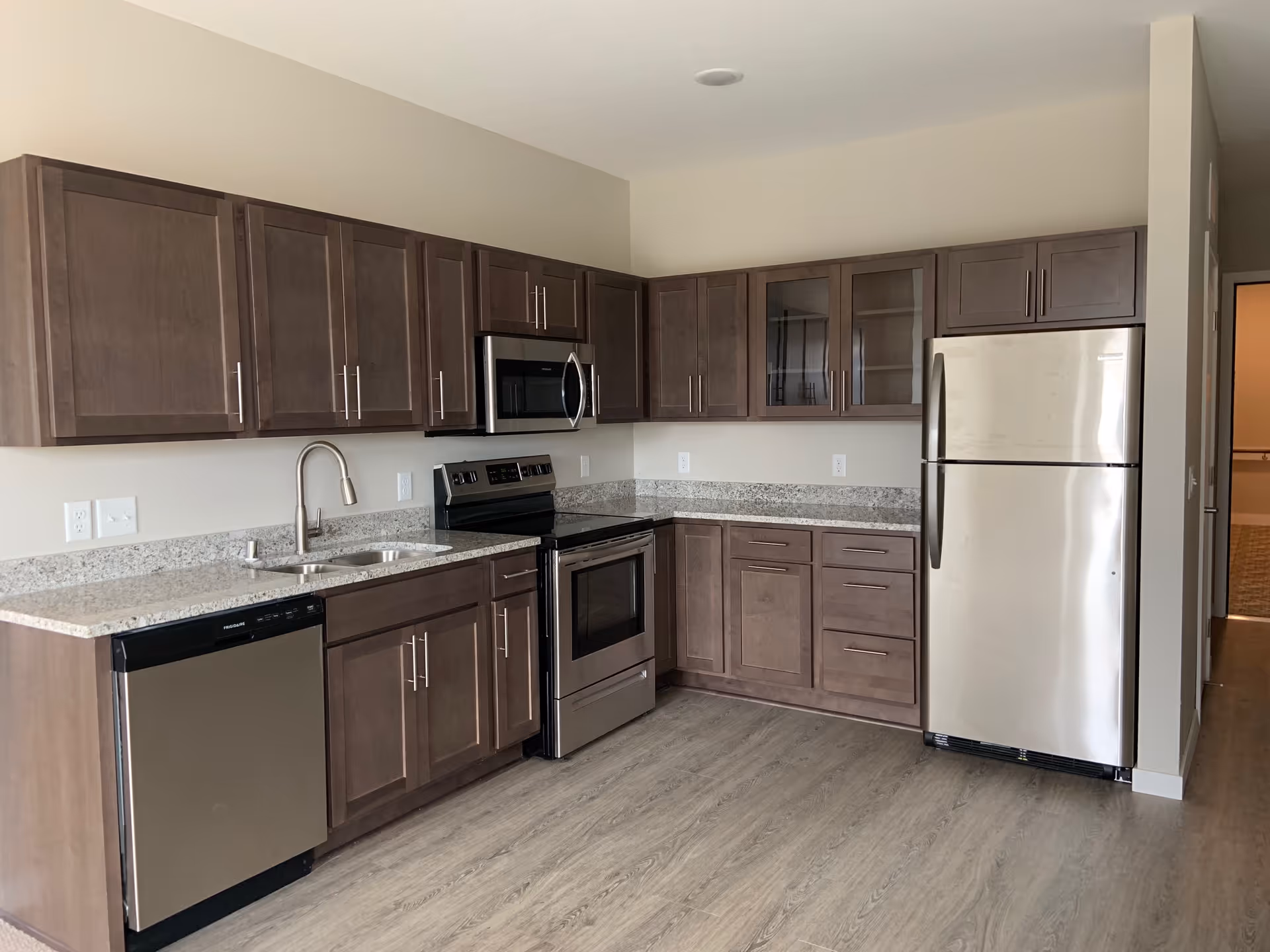 Modern kitchen with wooden cabinets, granite countertops, stainless steel appliances including a refrigerator, oven, microwave, and dishwasher, and wood-style flooring.