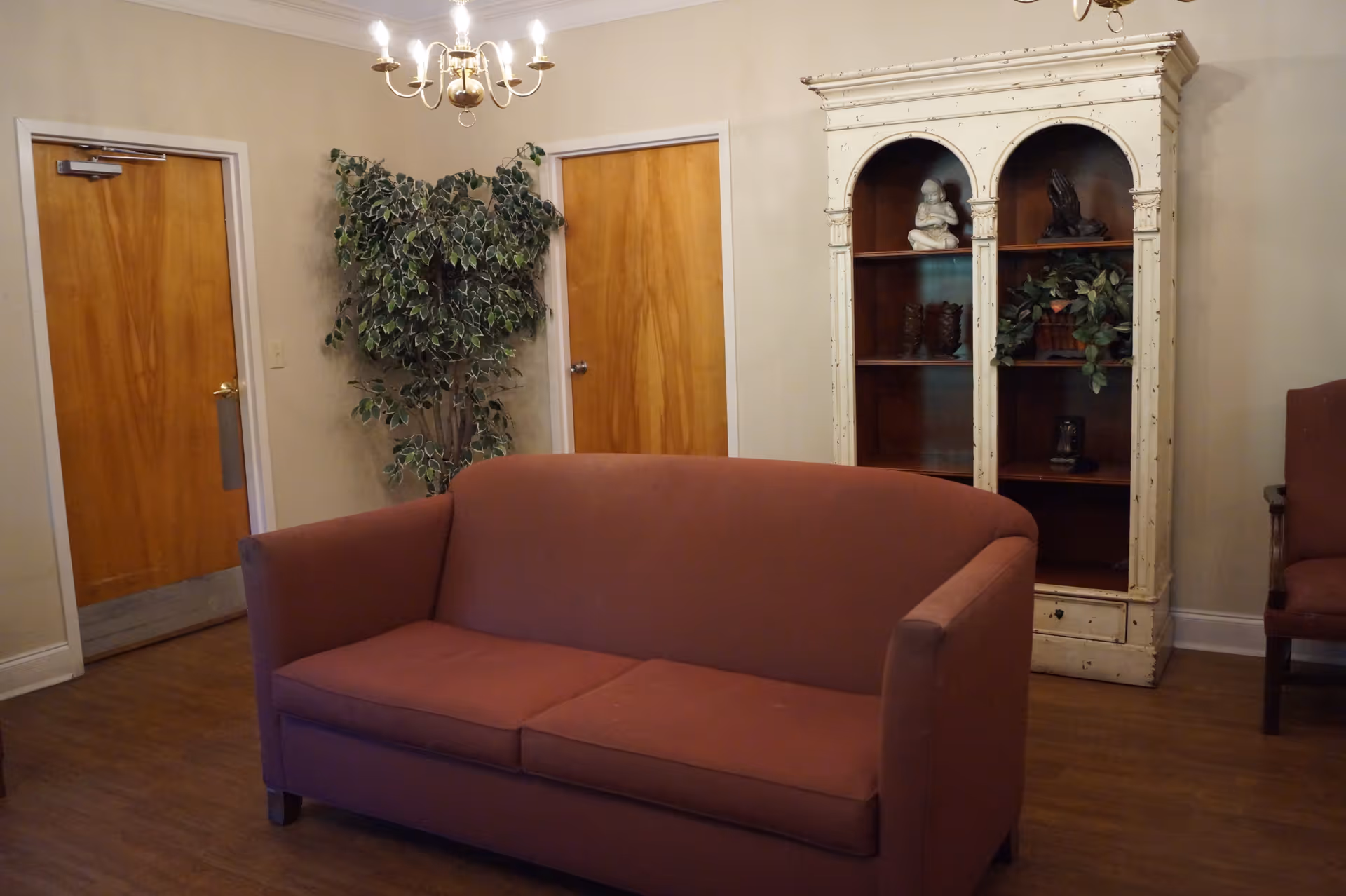 Small sitting room with a red sofa, wooden doors, a tall white display cabinet, potted plant, and chandelier.