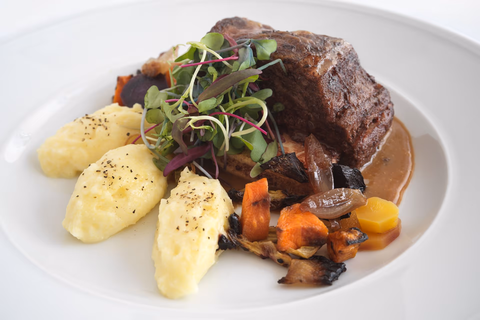 A plated gourmet meal featuring a piece of braised meat with brown sauce, three quenelles of mashed potatoes sprinkled with black pepper, roasted vegetables including carrots and onions, and a garnish of fresh microgreens on a white plate.