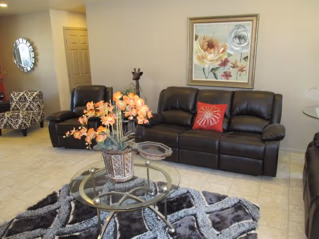 A living room with a black leather sofa and matching recliner chairs. There is a glass coffee table with a decorative flower arrangement on top, placed on a patterned area rug. A framed floral painting hangs on the beige wall behind the sofa, and a red decorative pillow is on the sofa. In the background, there is a patterned armchair and a round mirror on the wall near a doorway.