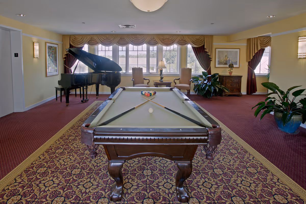 A recreational room with a pool table in the center, a grand piano to the left, two chairs and a small table near large windows with curtains, and several potted plants. The room has patterned carpet and yellow walls with framed artwork.