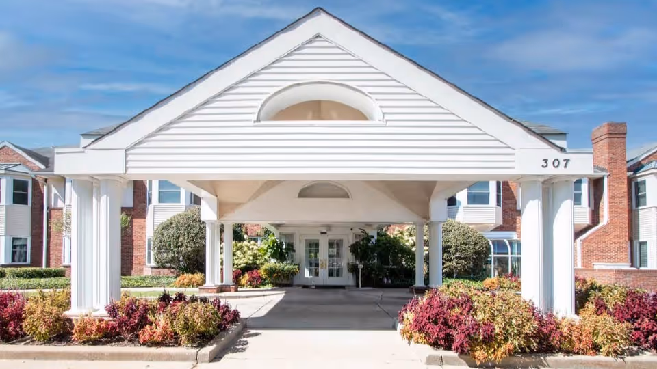 Front exterior view of a senior living facility named Brooking Park, featuring a white covered entrance with columns, landscaped bushes and plants, and a brick building under a blue sky.