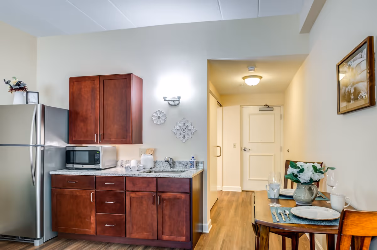 Interior view of a small kitchen and dining area in a senior living facility. The kitchen features dark wooden cabinets, a stainless steel refrigerator, a microwave, and a granite countertop with a sink. On the right, there is a wooden dining table set for two with plates, glasses, utensils, and a vase with white flowers. The background shows a hallway leading to a closed door, with light-colored walls and wood flooring throughout.
