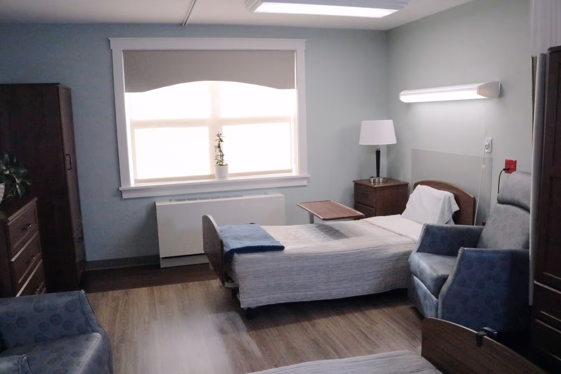 A simple, clean bedroom in a senior living facility with a single bed, a bedside table with a lamp, a reclining chair, a window with a blind, and wooden furniture including a dresser and a wardrobe.