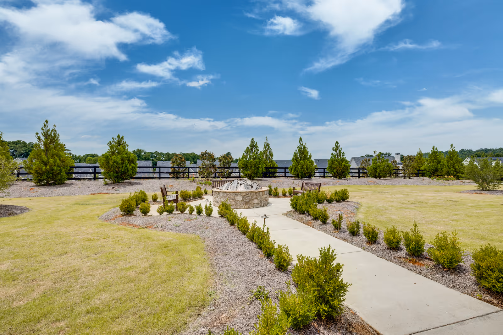 Landscaped outdoor courtyard with a central stone fire pit, benches, a paved walkway, and young shrubs under a blue sky.