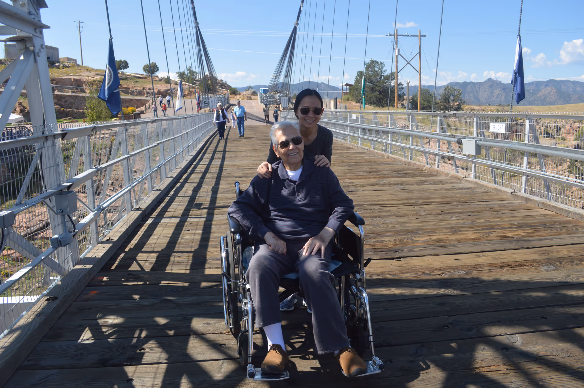 An elderly man in a wheelchair is smiling while being pushed by a woman wearing sunglasses on a wooden suspension bridge. Other people are walking in the background, and there are mountains and a clear blue sky visible.