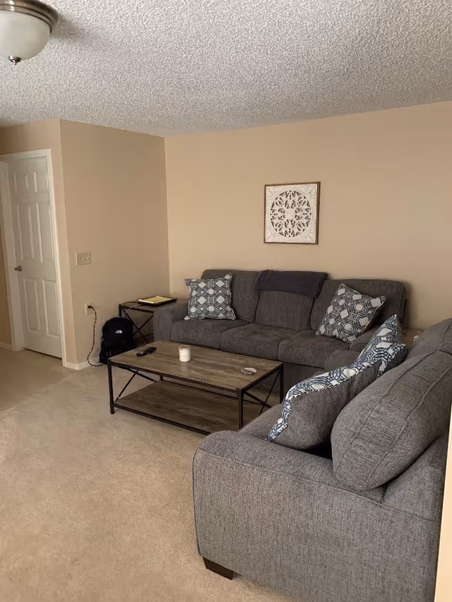 A living room with beige walls and carpet, featuring two gray sofas with patterned blue and white cushions. A wooden coffee table with a metal frame sits between the sofas, holding a candle and a remote control. A small side table with a notebook and a backpack is placed near a closed white door. A decorative framed wall art hangs above one of the sofas.