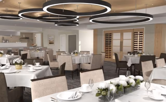 A spacious dining room with multiple round tables covered with white tablecloths, each set with plates, glasses, and cutlery. Beige upholstered chairs surround the tables. Modern circular ceiling lights hang above, and a wine storage area with wooden doors is visible in the background.