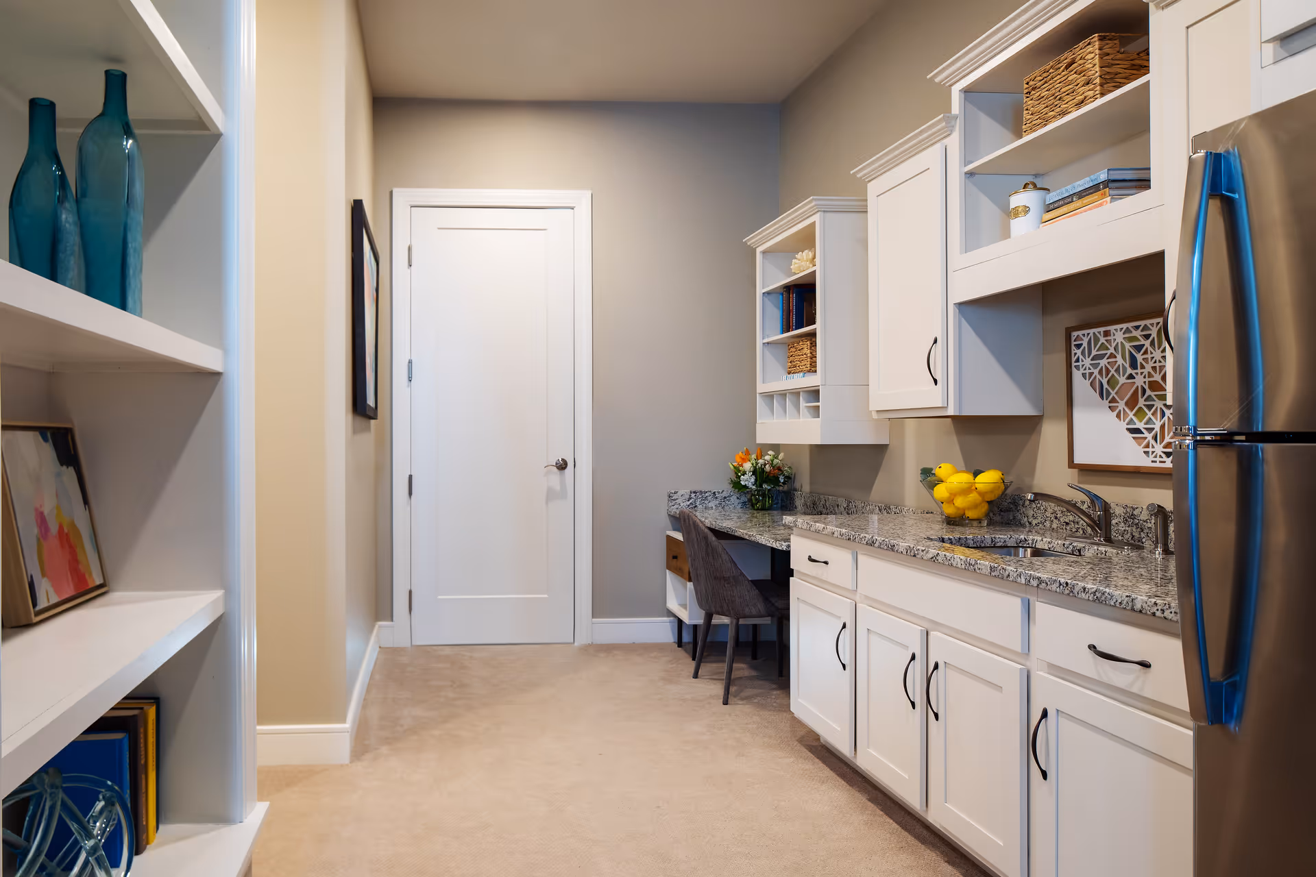 Interior view of a kitchen area in a senior living facility featuring white cabinets with black handles, a granite countertop with a sink, a stainless steel refrigerator, and a small desk area with a chair. Shelves on the left hold decorative blue glass bottles and framed artwork. The walls are painted beige and light gray, and the floor is carpeted.