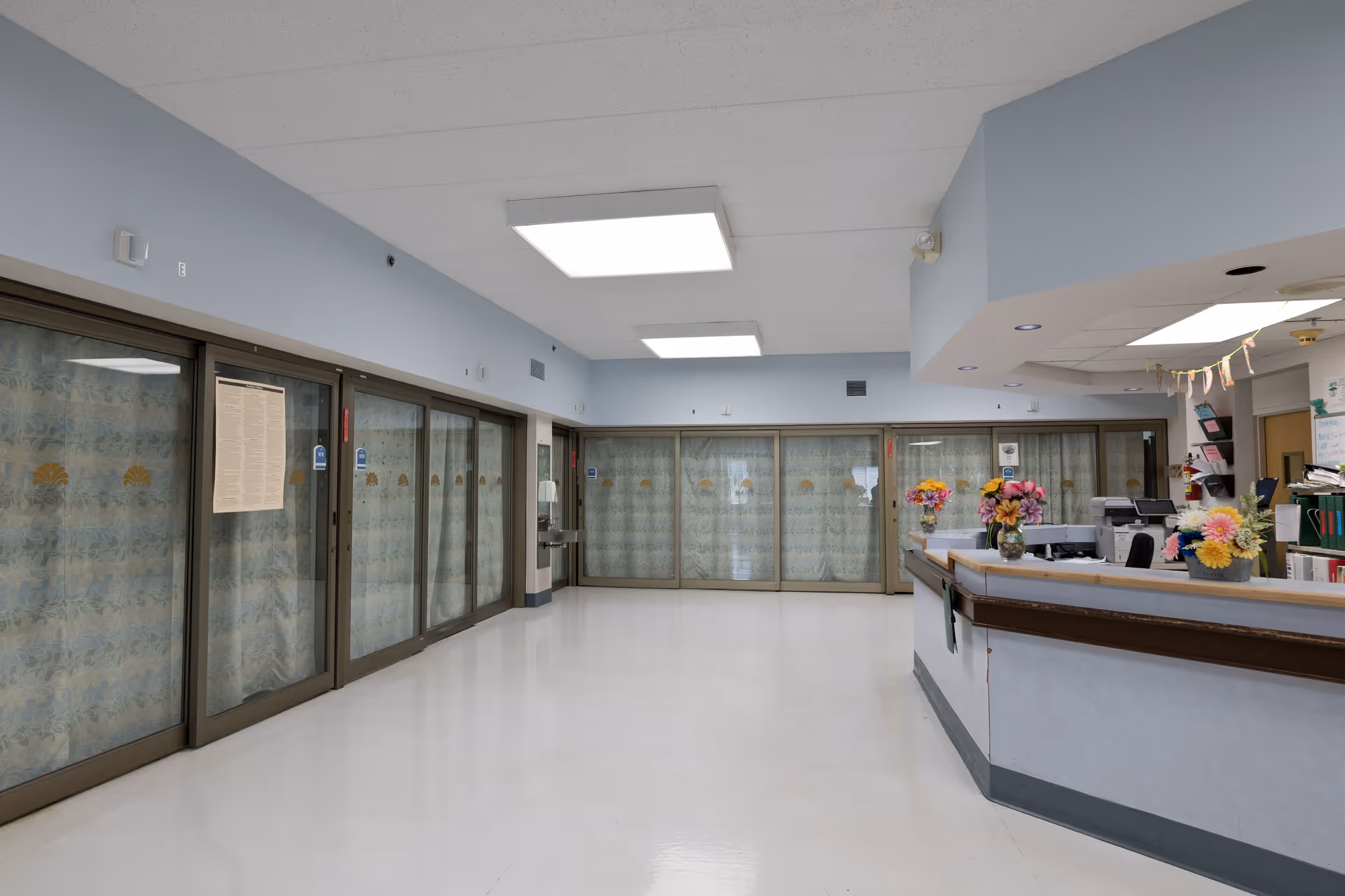 A clean, well-lit hallway in a healthcare facility with large sliding glass doors covered by patterned curtains on the left and at the end. On the right side, there is a reception desk decorated with colorful flower arrangements and office supplies. The ceiling has rectangular fluorescent lights, and the floor is shiny and white.