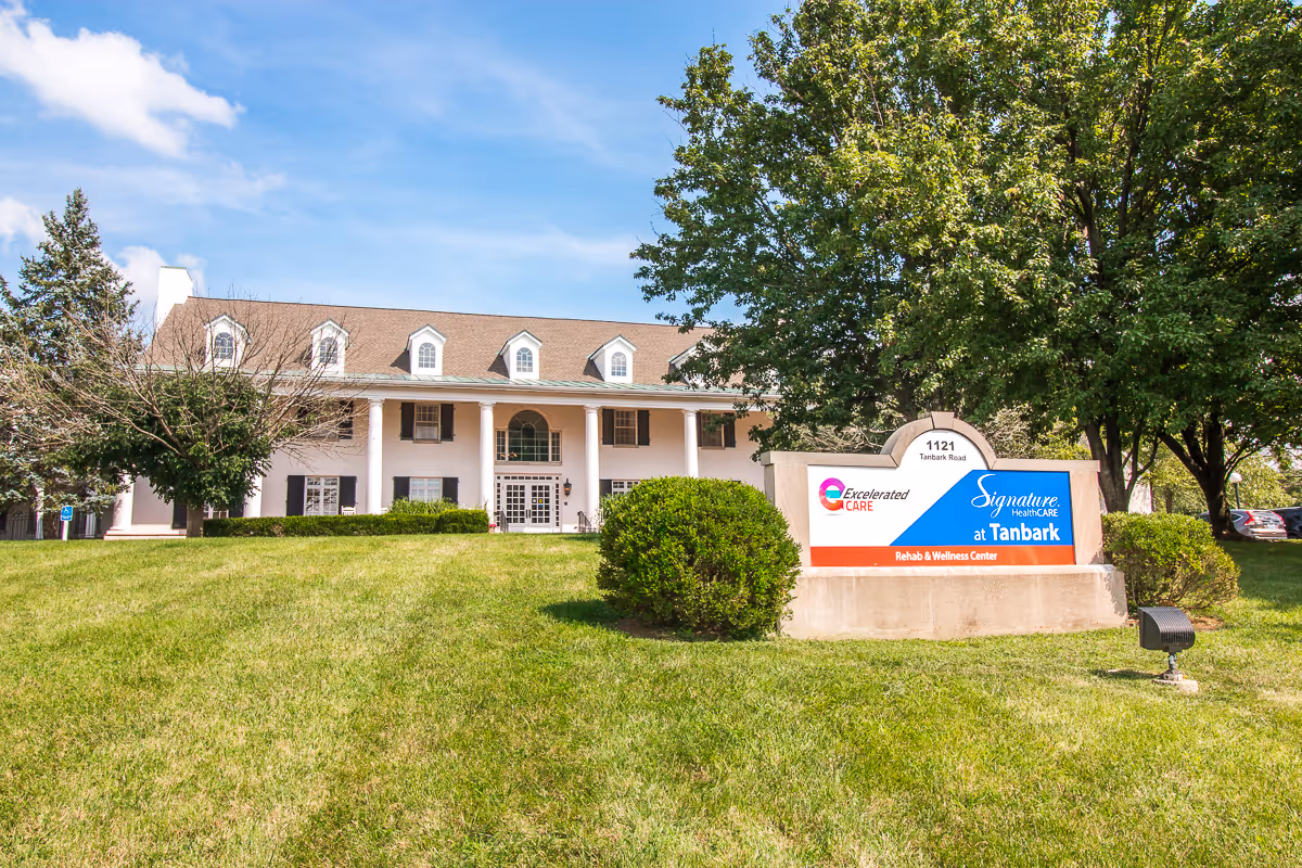 Exterior view of Signature HealthCARE at Tanbark Rehab & Wellness Center, a large white building with multiple windows and a brown roof. The building is surrounded by green grass, trees, and bushes. A sign in front displays the facility's name and address, 1121 Tanbark Road.