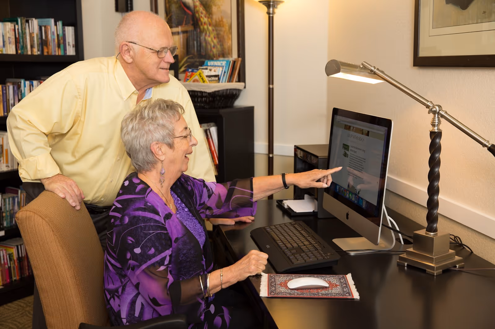 An elderly woman seated at a desk using a desktop computer, pointing at the screen, while an elderly man stands beside her looking at the screen. The room has bookshelves filled with books and a floor lamp providing light.