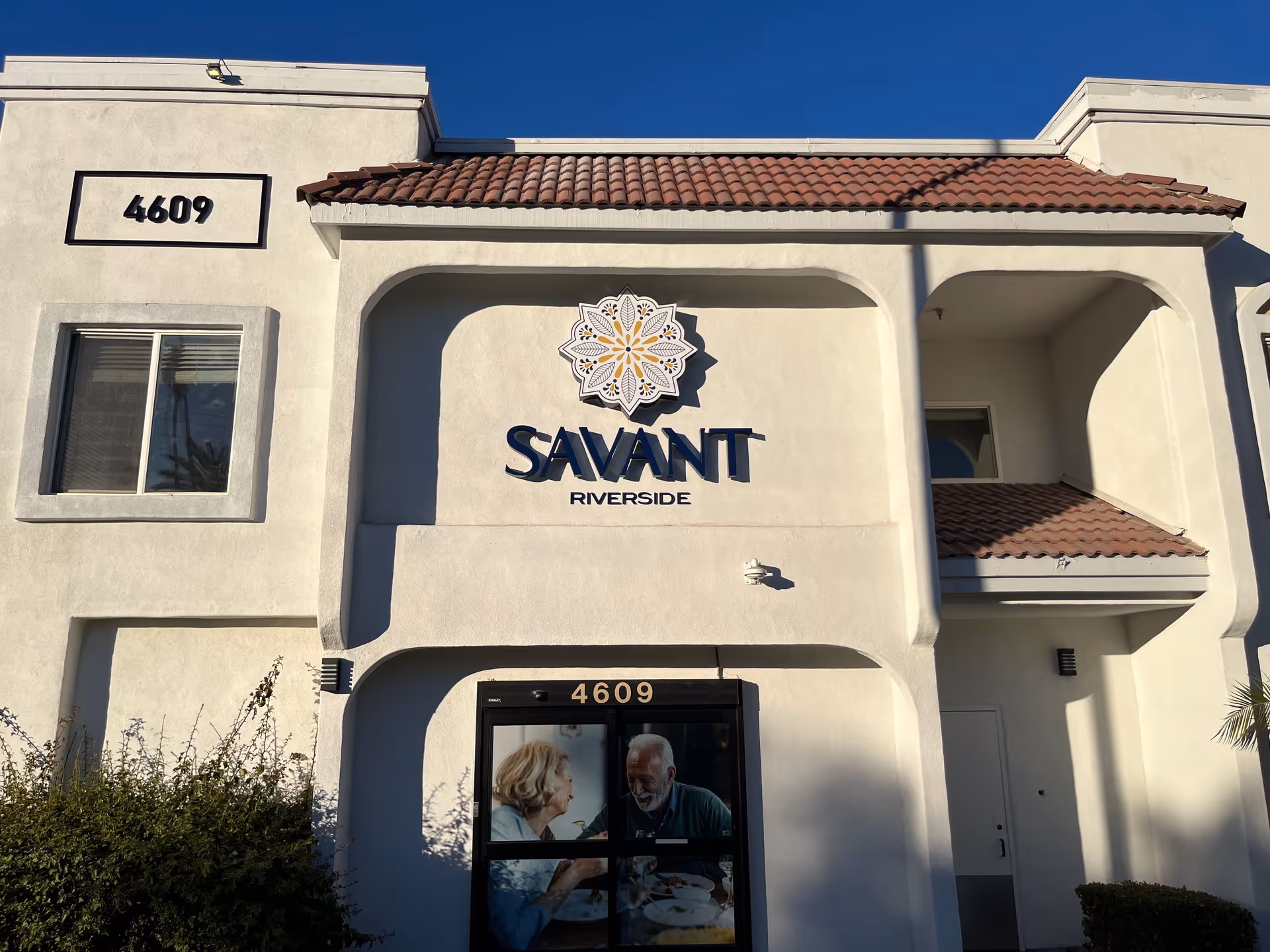 Exterior front view of a building with white walls and a red tile roof. The building has the address number 4609 displayed twice, once on the wall and once above the entrance door. Above the entrance is a sign that reads 'SAVANT RIVERSIDE' with a decorative emblem above the text. There is a window to the left of the entrance and some bushes in front of the building. The entrance door has an image of an elderly man and woman smiling and interacting.