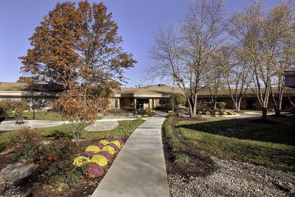 A paved walkway leads through a landscaped garden area with colorful flowers, bushes, and trees with autumn foliage toward the entrance of a single-story brick building under a clear blue sky.