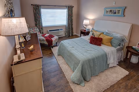 A cozy bedroom with a neatly made bed featuring a light blue quilt and multiple pillows in red, yellow, and blue. There is a wooden dresser with a lamp and decorative items on the left side, and a small round bedside table with a lamp on the right. A cushioned chair with a red throw blanket is positioned near a window with floral curtains. The floor is wooden with a light-colored rug under the bed.