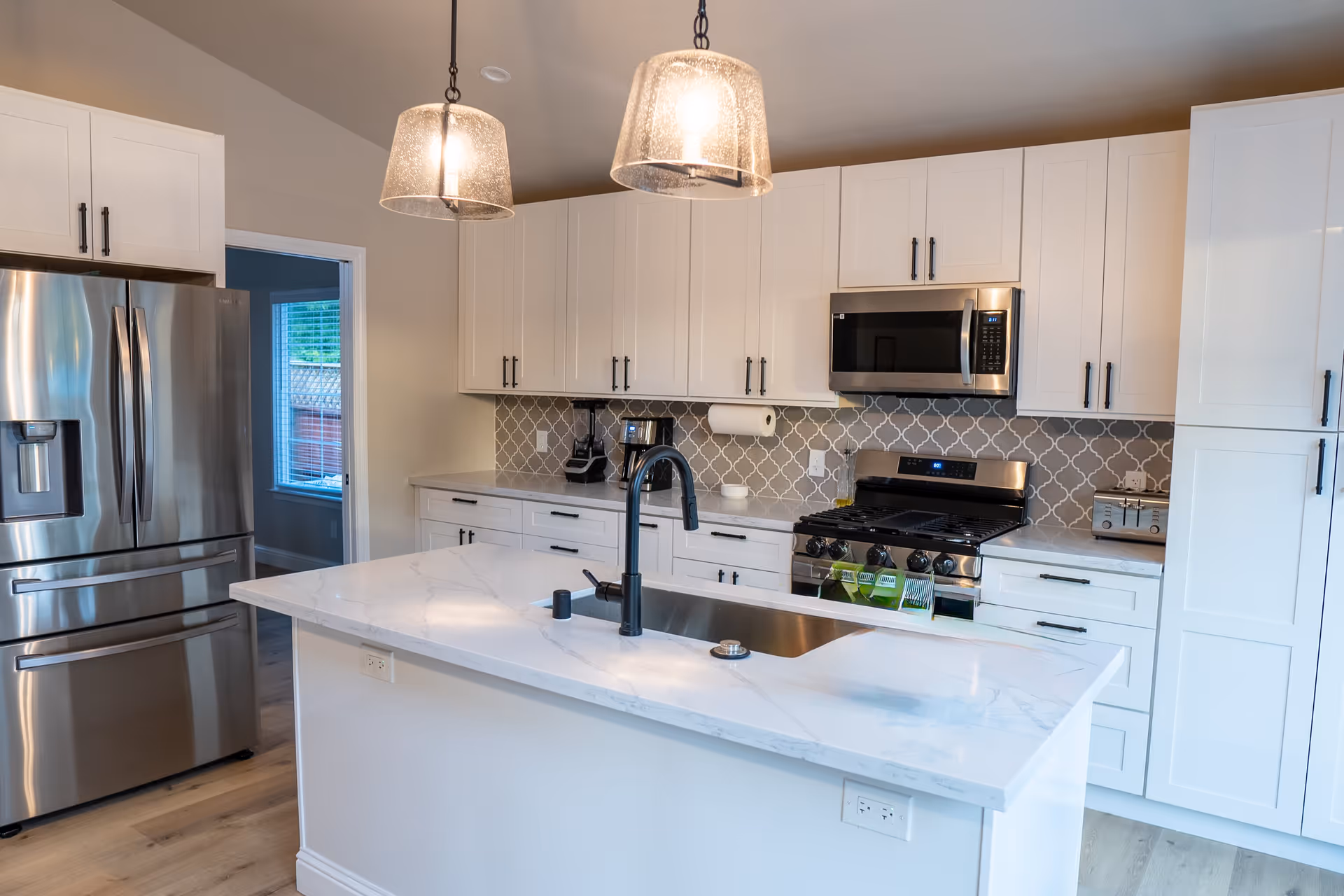 Modern kitchen with white cabinets, a marble countertop island with a black faucet and sink, stainless steel refrigerator, microwave, and stove. Two pendant lights hang above the island, and a patterned backsplash is visible behind the stove.