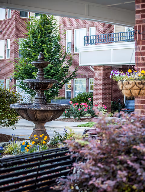 Outdoor courtyard area at Preston Pointe featuring a multi-tiered stone fountain surrounded by colorful flowers and greenery, with a brick building and windows in the background. There is a black metal bench in the foreground and a hanging basket with purple and yellow flowers on the right side.