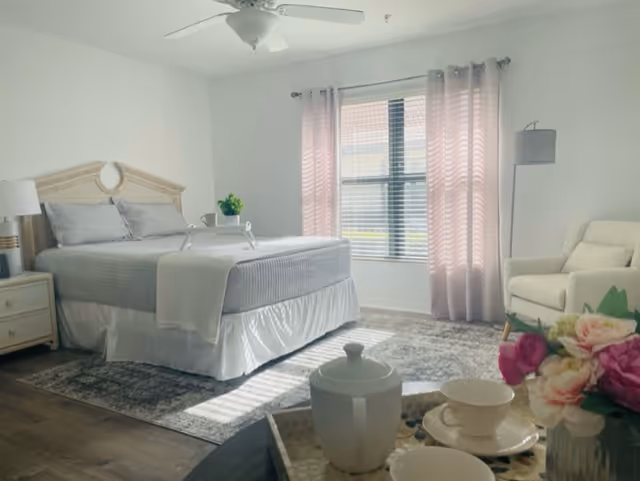 Sunlit furnished bedroom with a bed, nightstand, armchair, and a tray with a tea set and flowers in the foreground.