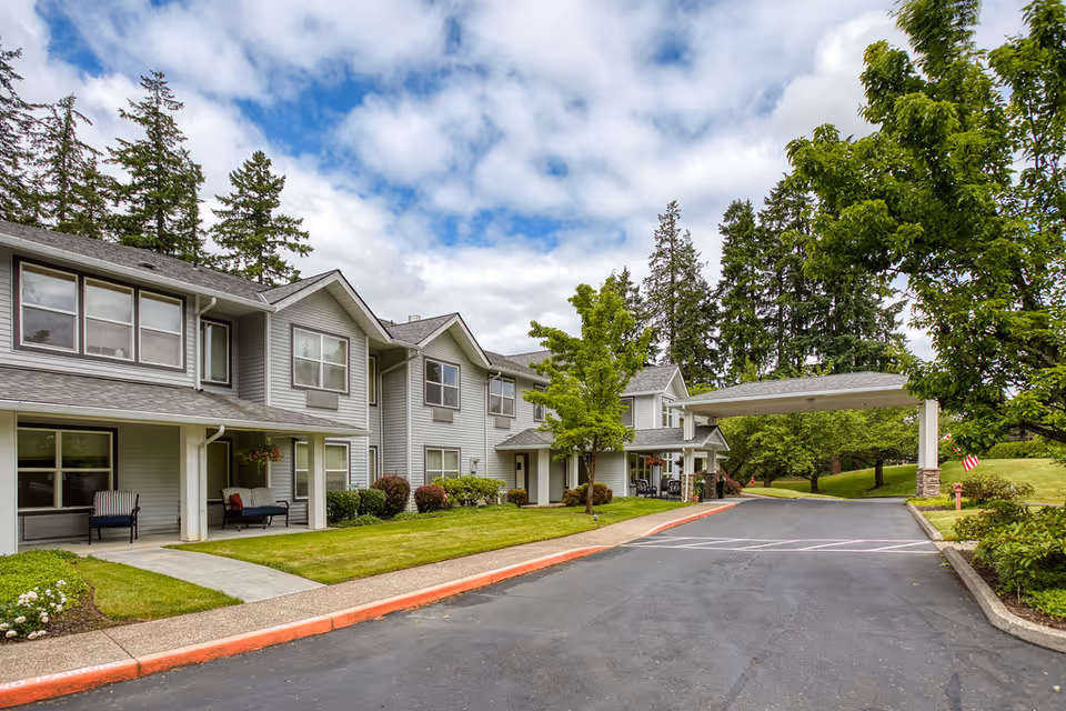 Exterior view of a two-story assisted living facility with gray siding, multiple windows, and a covered entrance. The building is surrounded by well-maintained green lawns, trees, and shrubs under a partly cloudy sky.
